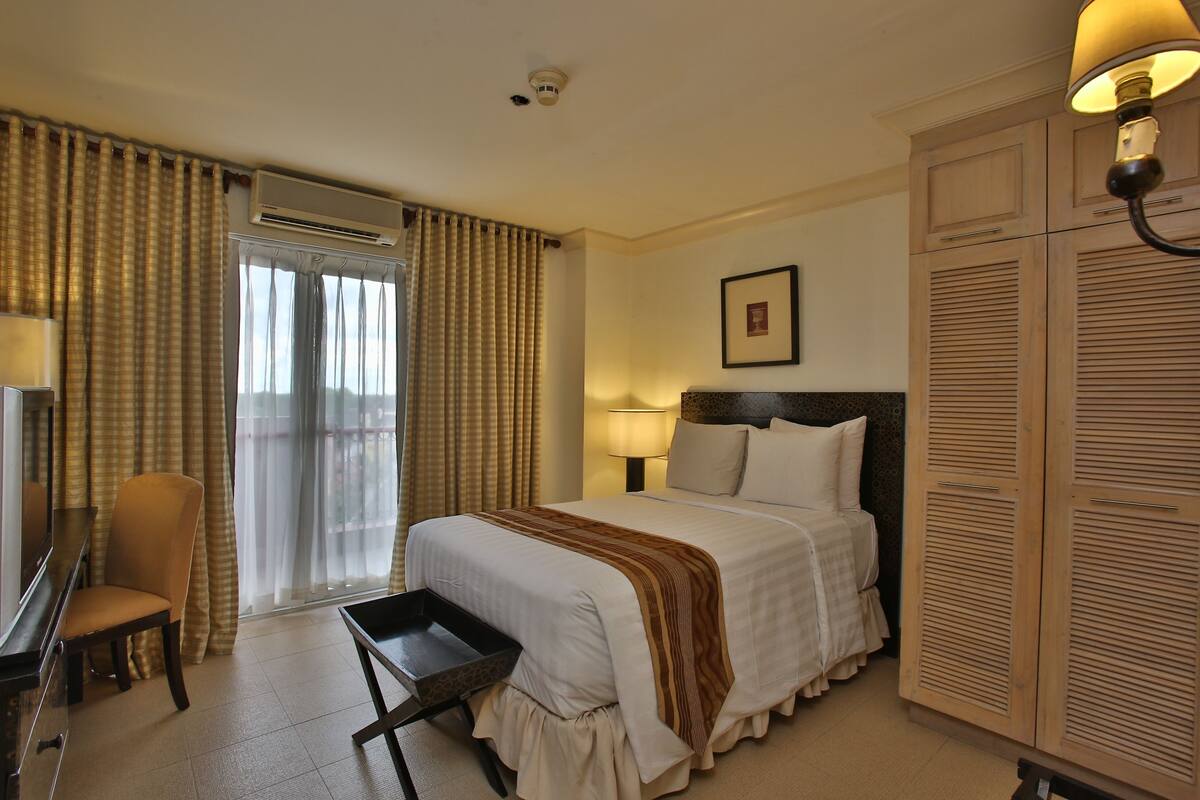 A hotel room with a bed, a desk, a chair, a wardrobe, and a window with curtains.