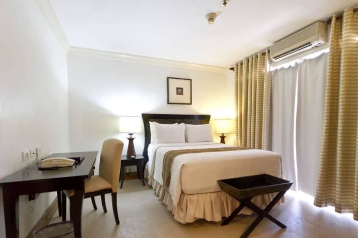 Una habitación de hotel con cama, mesita de noche, silla, escritorio y aire acondicionado.
