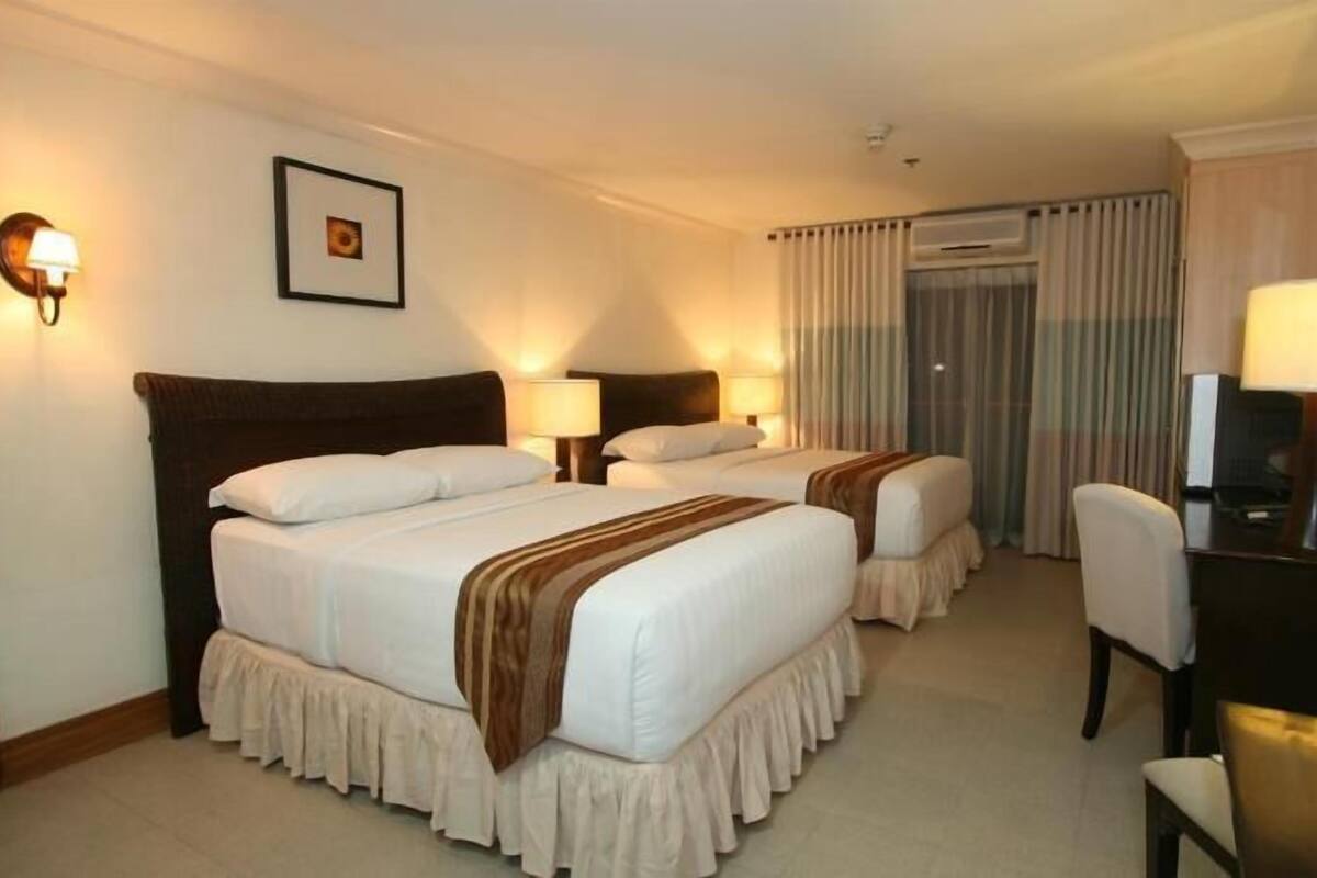 Una habitación de hotel con dos camas, un escritorio con silla, televisión y ventana con cortinas.
