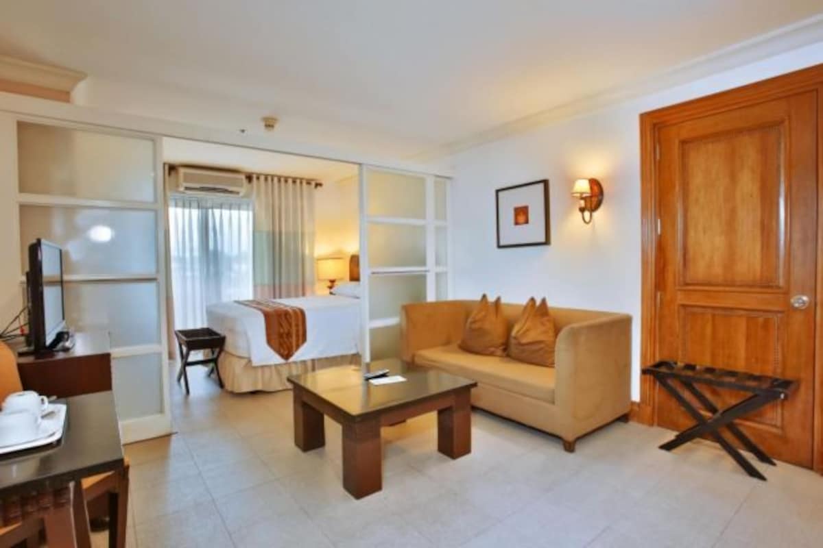 A hotel room with a bed, a sofa, a coffee table, a desk with a chair, a TV, and a wooden door.