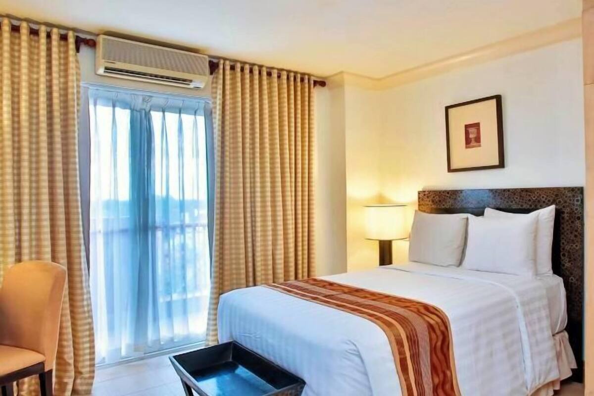 A hotel room with a bed, a chair, a window with curtains, and a framed picture on the wall.
