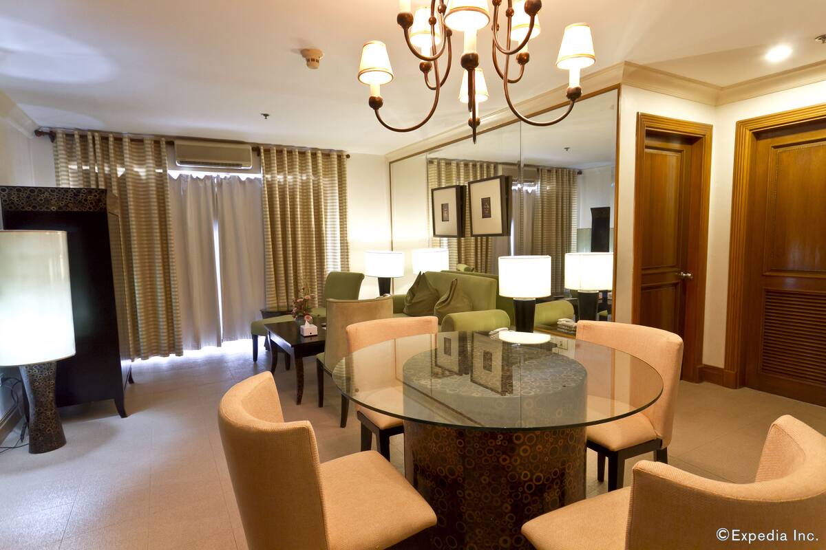 A hotel room with a round glass table surrounded by four chairs, a chandelier, a large mirror, and a television on a stand.
