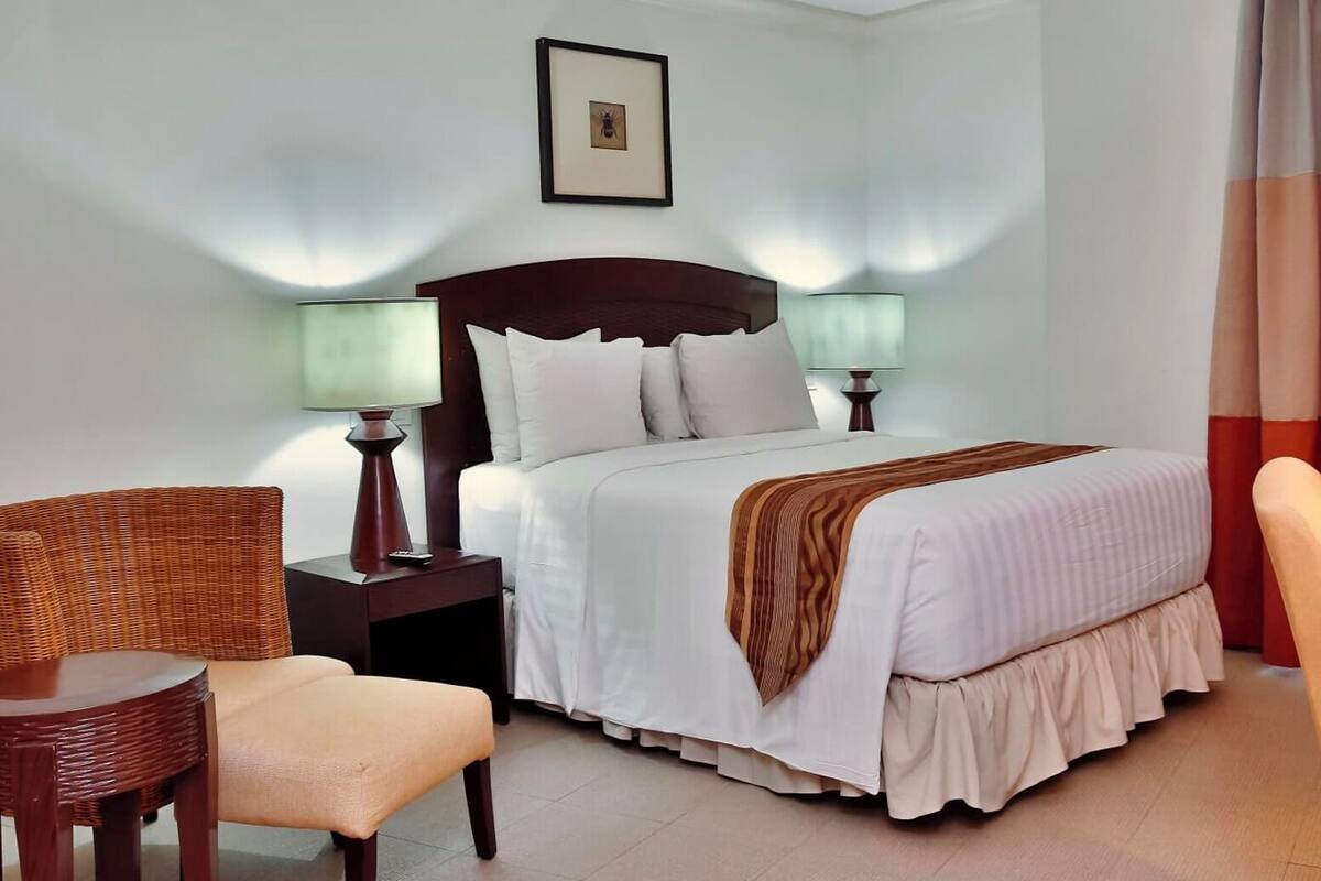 A hotel room with a bed, two bedside lamps, a chair, a small table, and a picture on the wall.