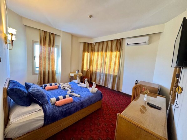 A hotel room with a large bed, a desk, a chair, and an air conditioning unit.