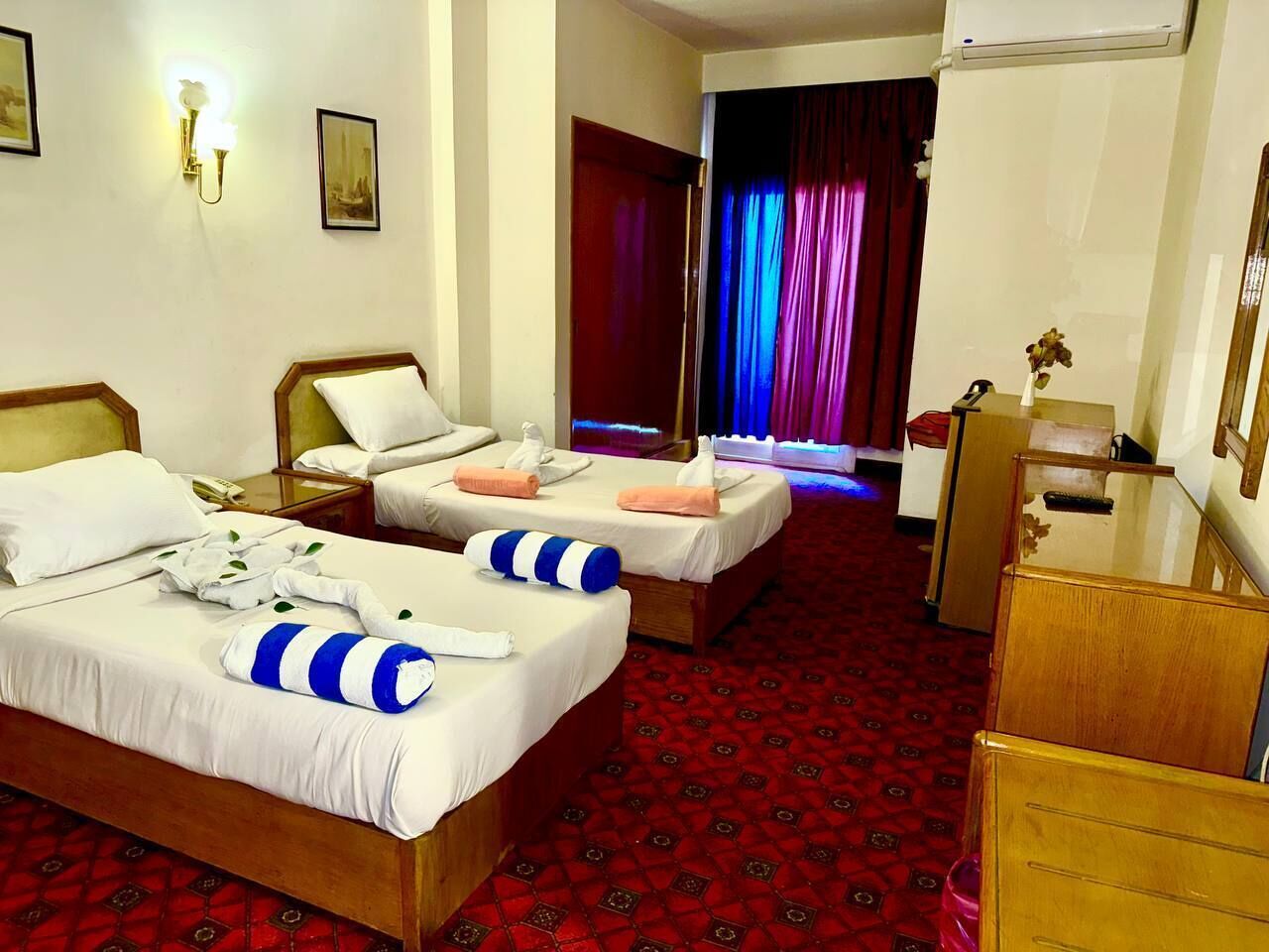 A hotel room with two beds, a wooden dresser, a desk, and a red patterned carpet.