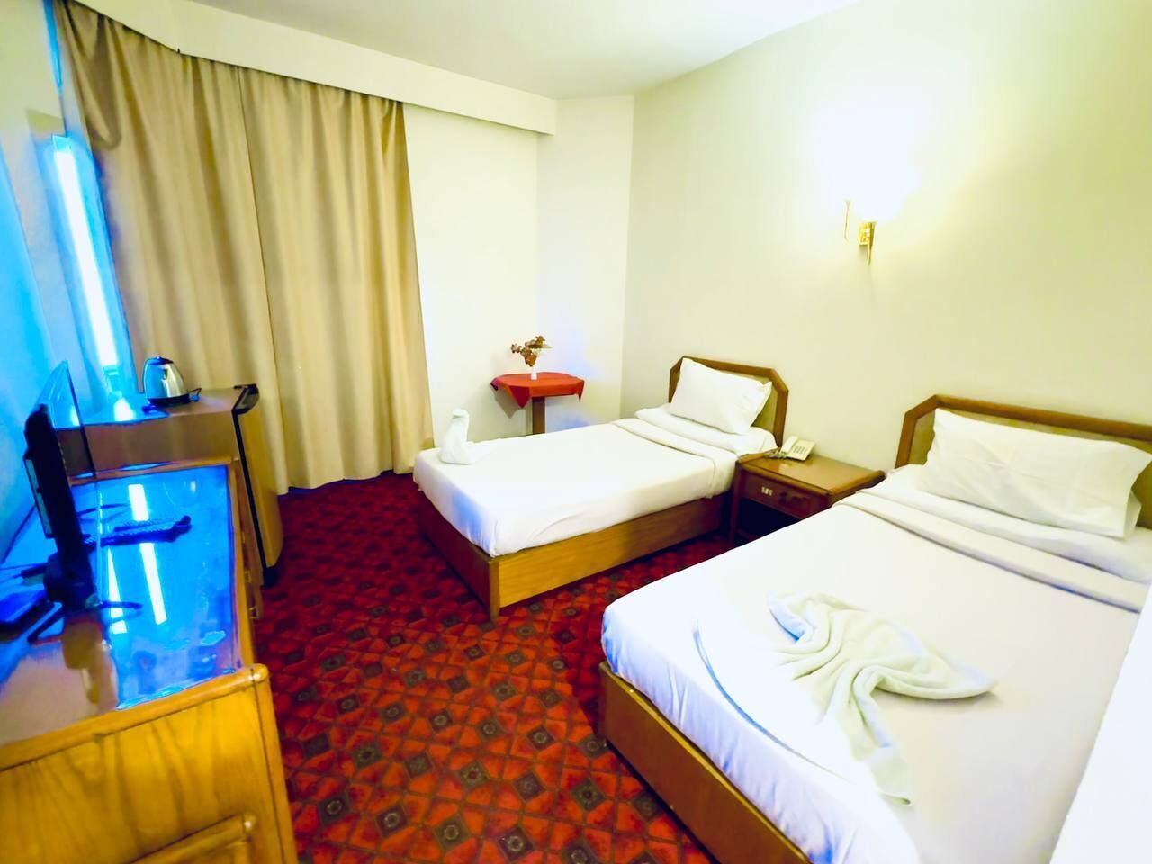A hotel room with two beds, a desk with a computer, a small table with a vase, and a red patterned carpet.