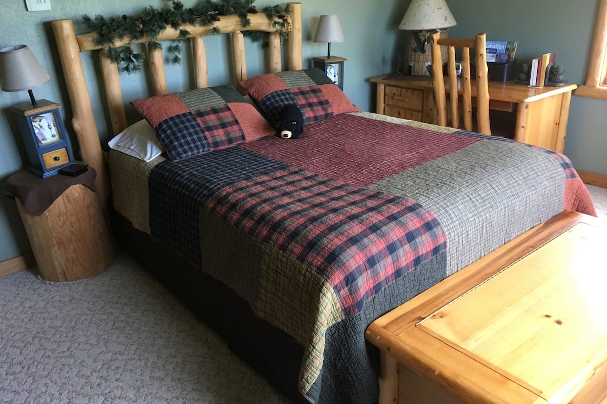 A bed with a plaid bedspread, wooden headboard with greenery, two bedside tables with lamps, and a wooden nightstand.