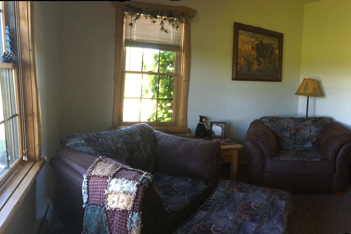 A cozy living room with a sofa, armchair, and a large window with blinds. There's a door leading to another room and a bathtub visible in the corner.