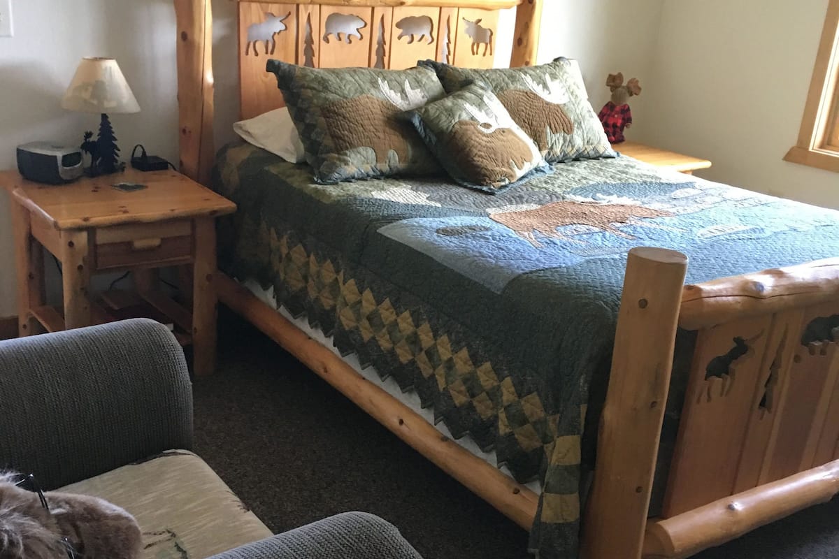 A wooden bed with a quilt, a nightstand with a lamp, and a chair with a blanket.