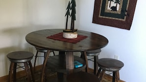 A round dining table with four wooden stools, a central lamp, and a rug with a forest pattern.