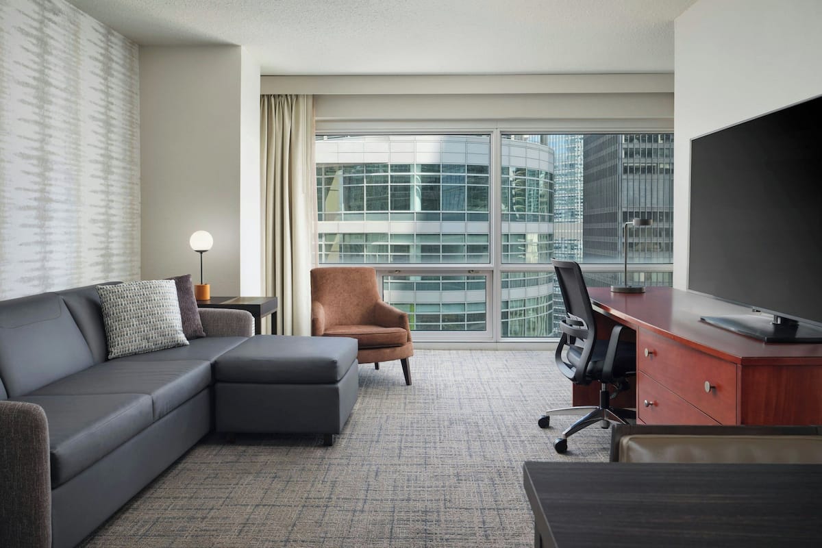 A modern hotel room with a grey sofa, a brown armchair, a desk with a computer, and a large window offering a city view.