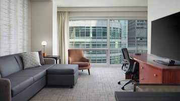 Quarto de hotel moderno com um sofá cinzento, uma poltrona castanha, uma escrivaninha com um computador e uma janela ampla com vista para a cidade.