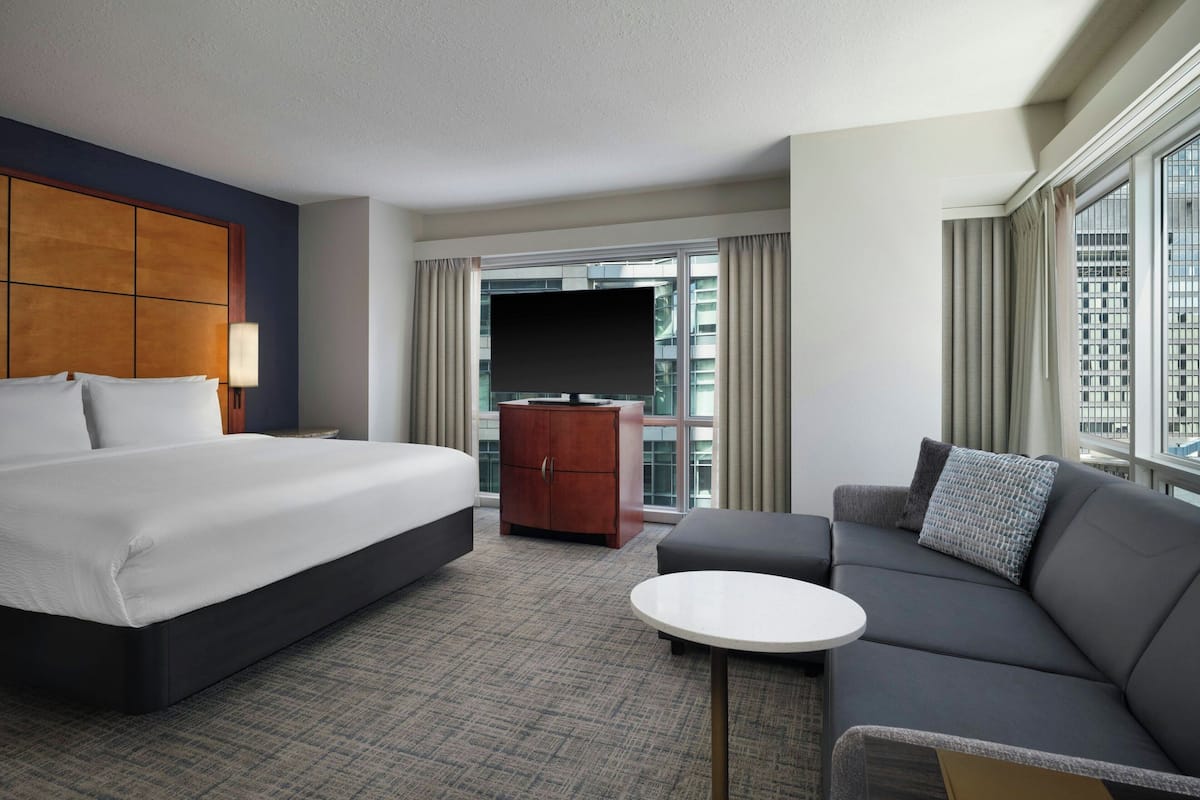 A hotel room with a large bed, a sofa, a small table, a TV, and a view of the city through the window.