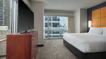 A hotel room with a large bed, a flat-screen TV on a wooden dresser, and a city view through large windows.