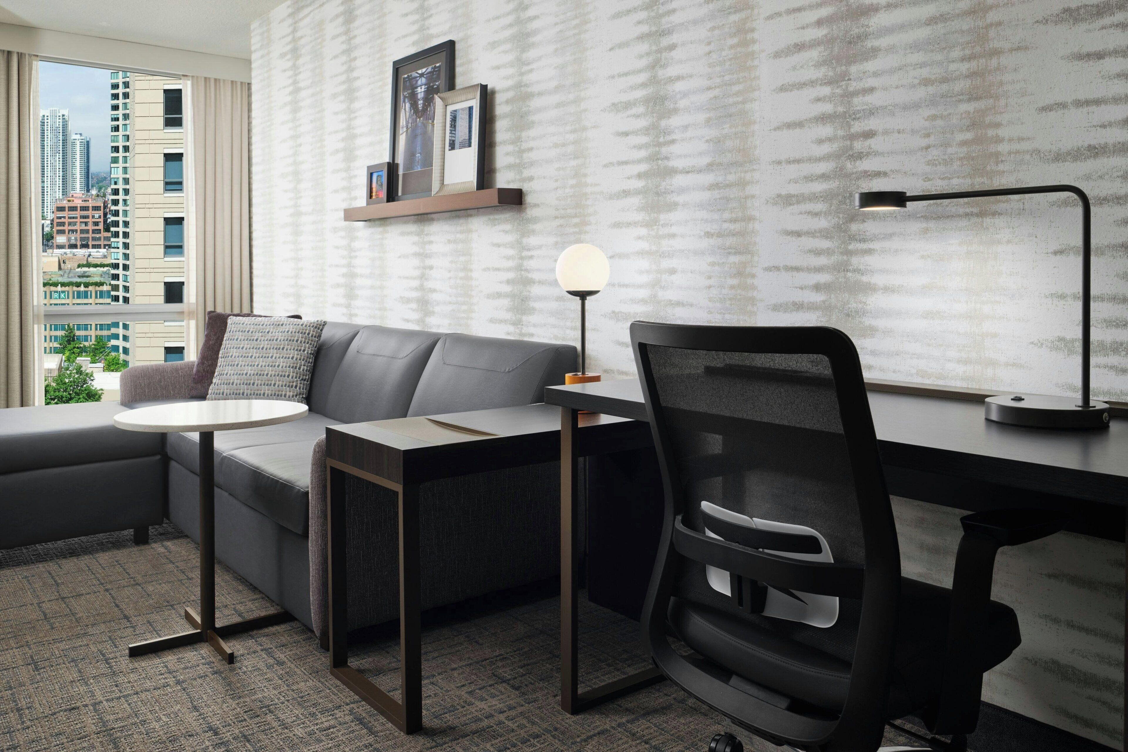 Una habitación de hotel moderna con un sofá gris, una pequeña mesa redonda, una silla de oficina negra, un escritorio y vistas de edificios altos a través de ventanas grandes.