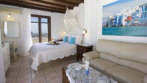 A bedroom with a bed, a sofa, a table with a vase, and a painting of a coastal scene.