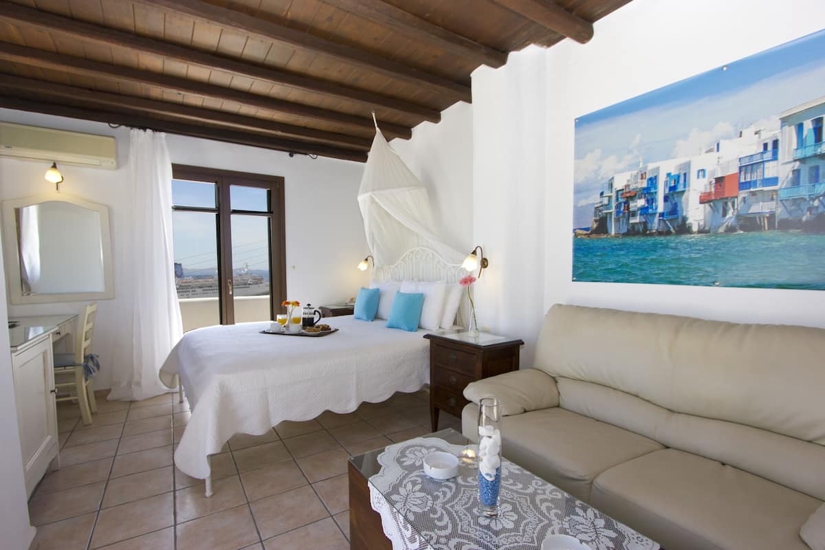A bedroom with a bed, a sofa, a table with a vase, and a painting of a coastal scene.