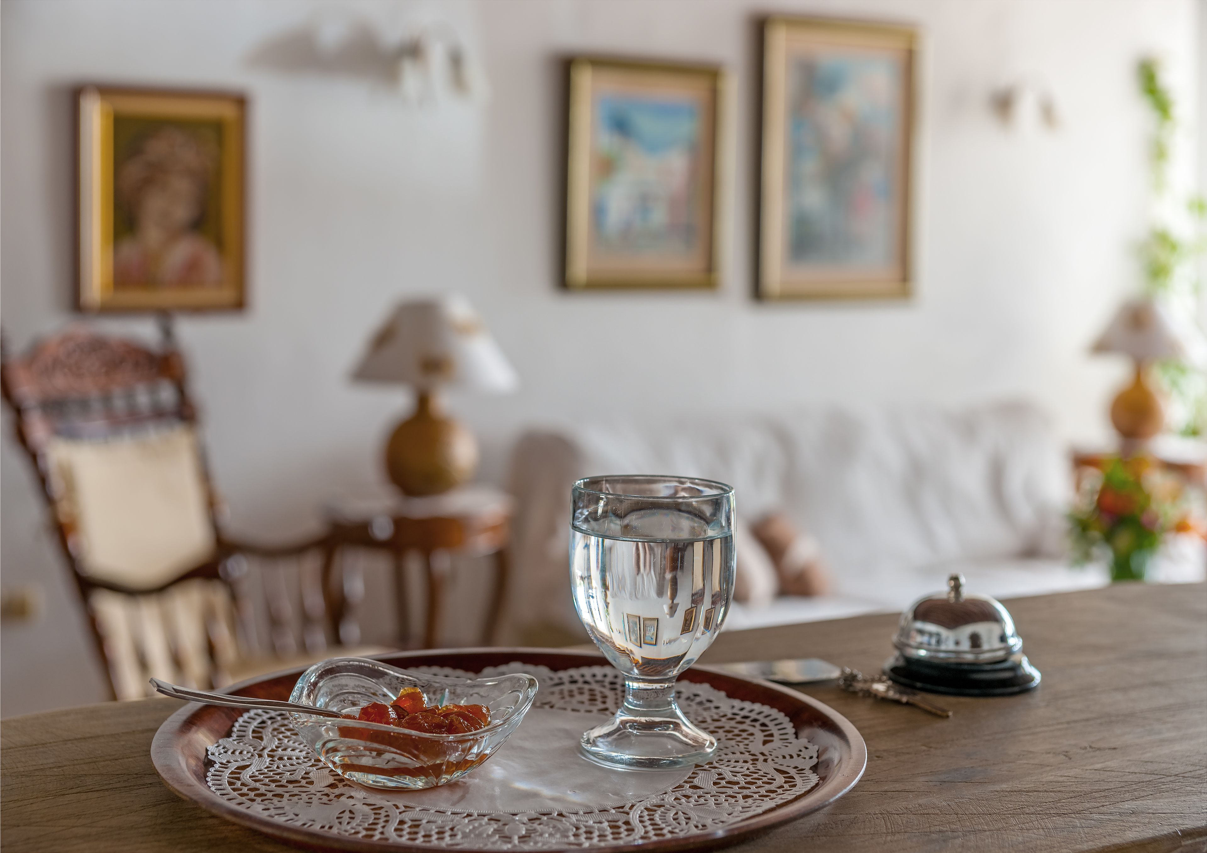 A dining table with a clear glass, a small bowl of fruit, and a decorative plate. There are framed pictures on the wall and a lamp on the side.
