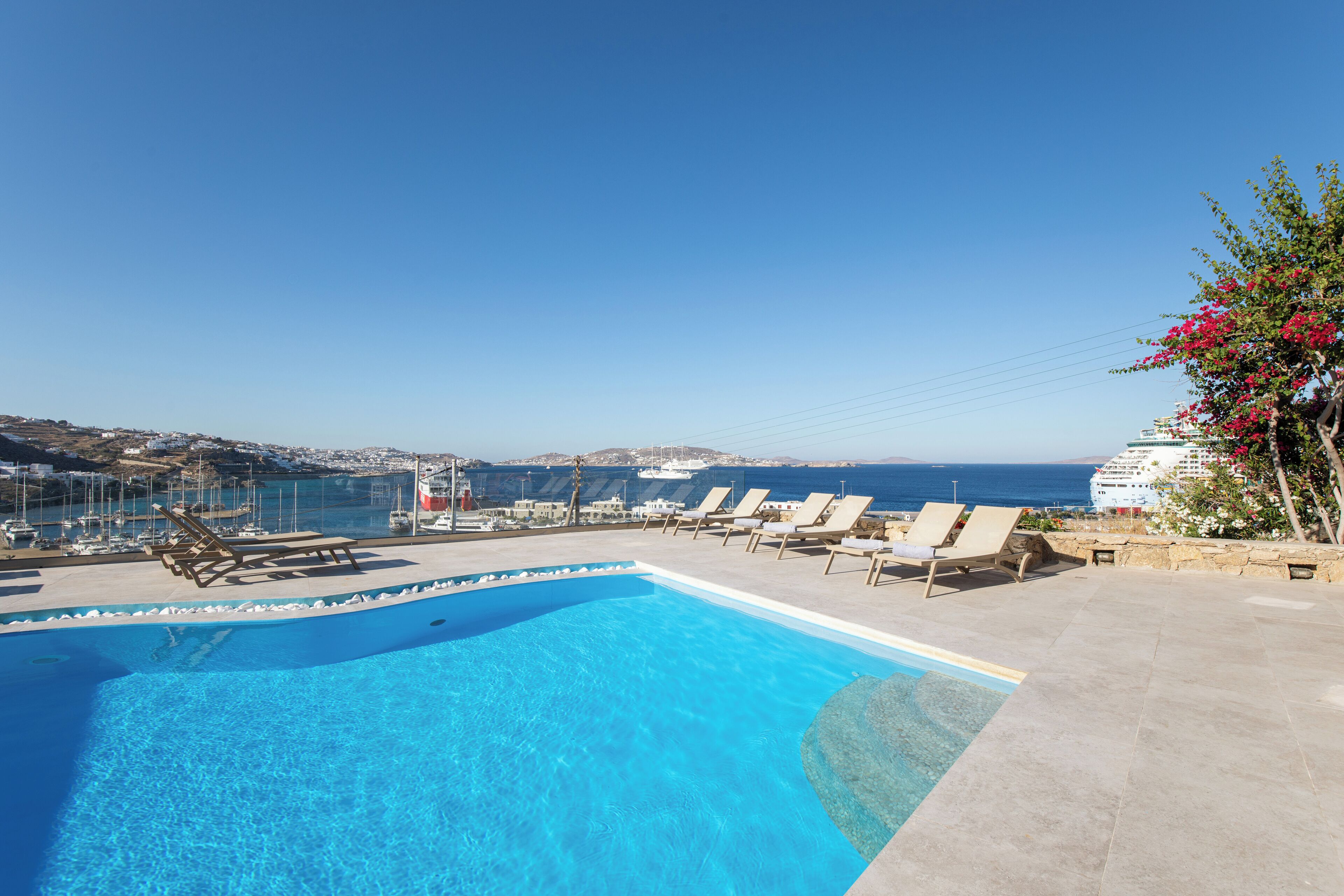 A clear blue swimming pool with lounge chairs and a view of the sea and boats.