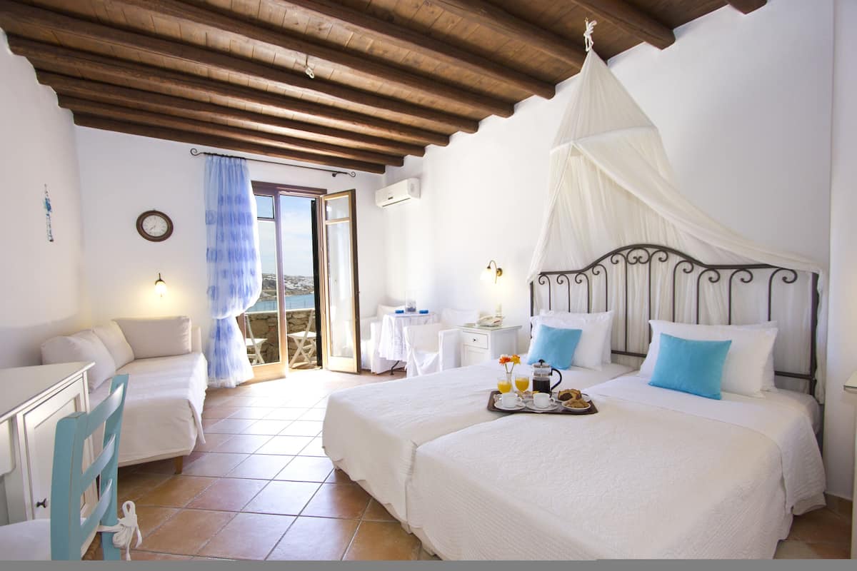 A bedroom with two beds, a dining table, and a chair. There is a balcony with a view.