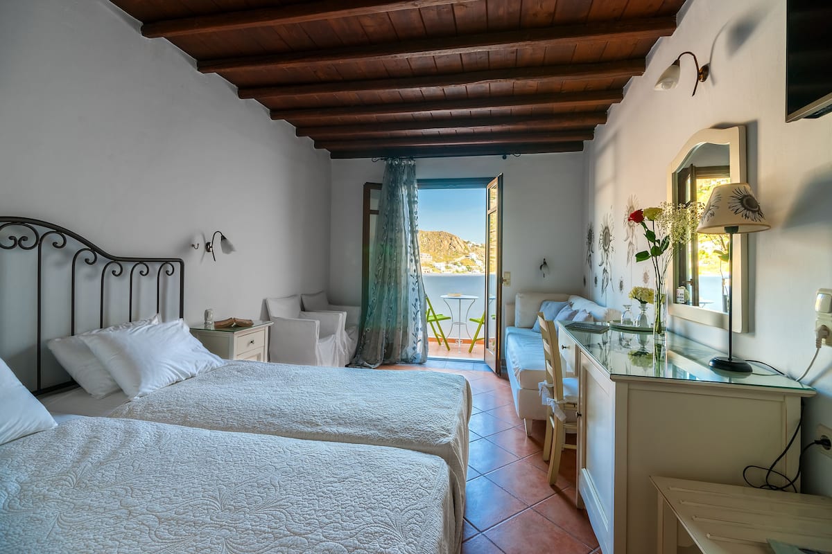 A bedroom with two beds, a desk, a chair, and a balcony with a view of the hills.