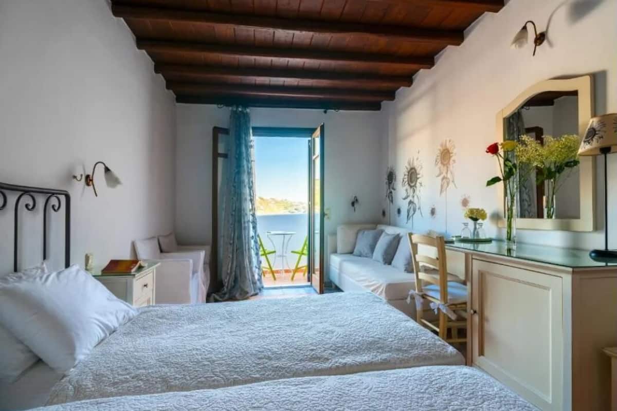 A bedroom with two beds, a wooden ceiling, a balcony with a table and chairs, and a view of the sea.