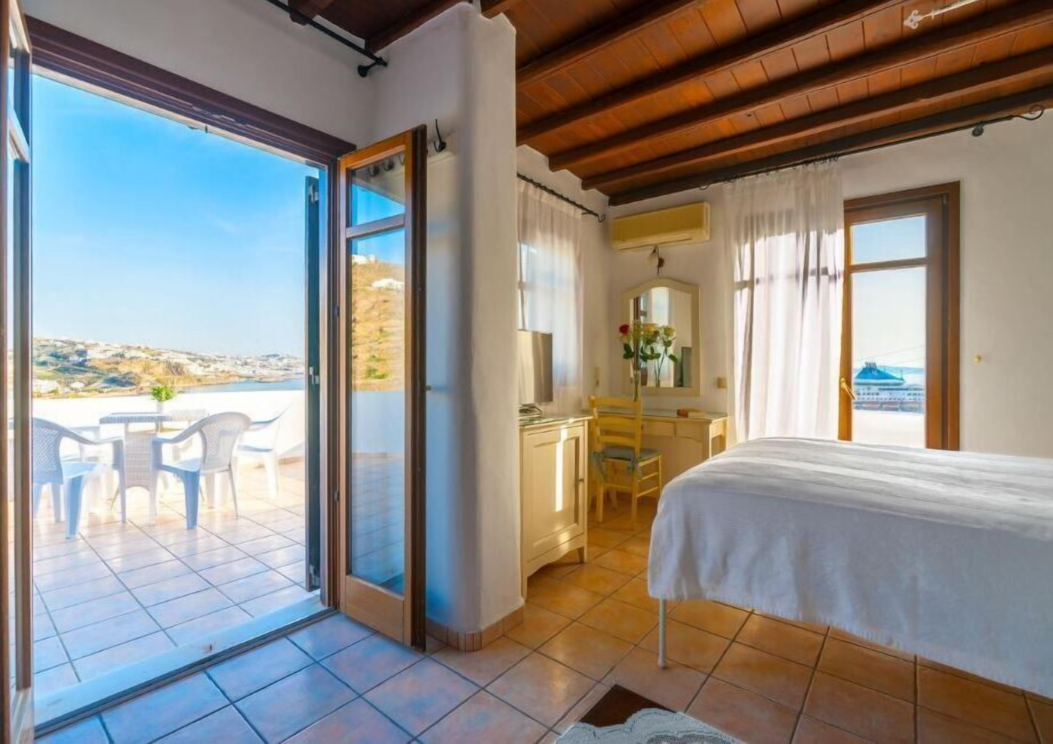 Une chambre avec un lit, un bureau et une chaise, un balcon avec une table et des chaises, et une vue sur la mer.
