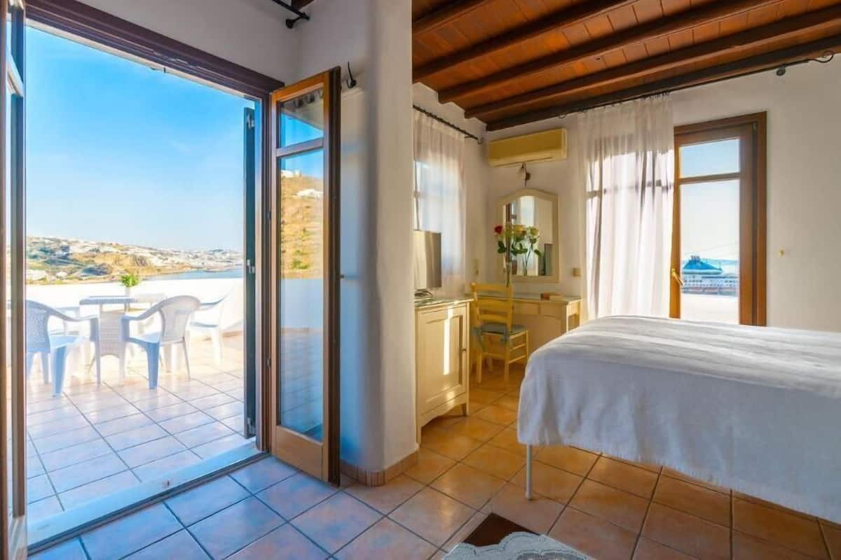 Une chambre avec un lit, un bureau et une chaise, un balcon avec une table et des chaises, et une vue sur la mer.
