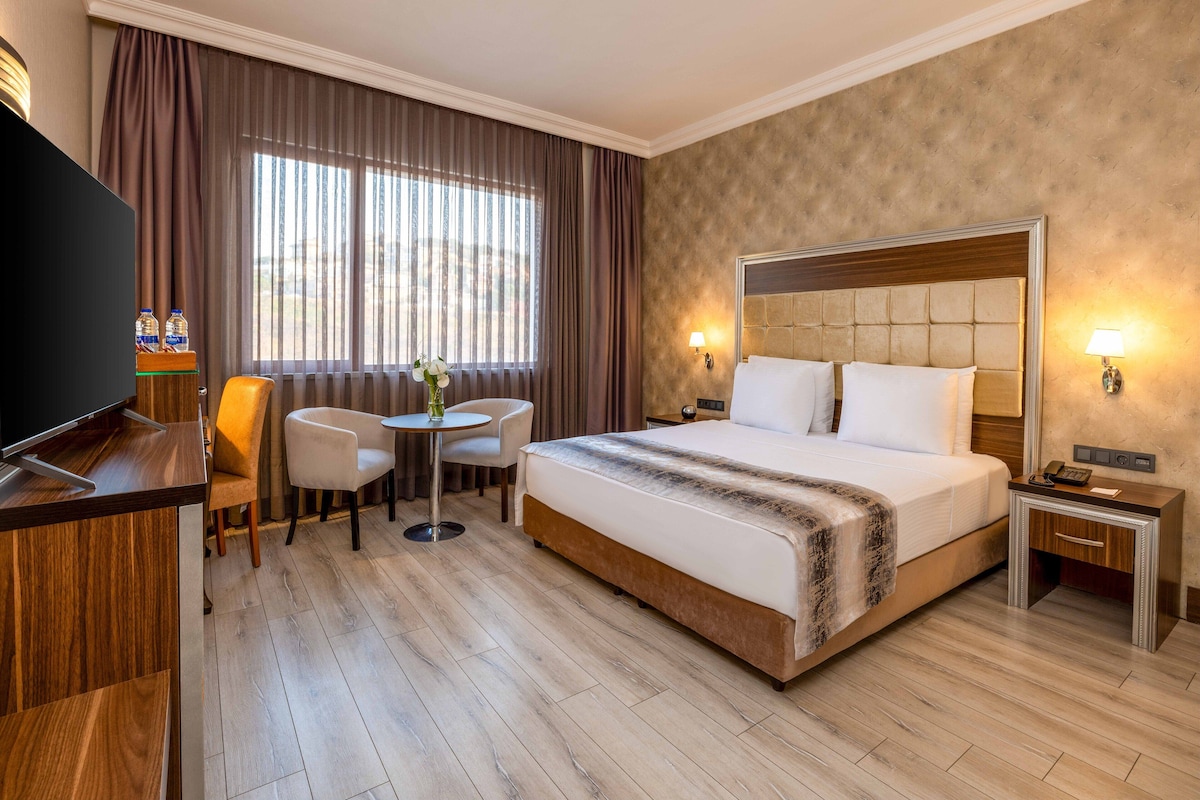 A hotel room with a large bed, a desk with a TV, two chairs, a small table, and a window with curtains.