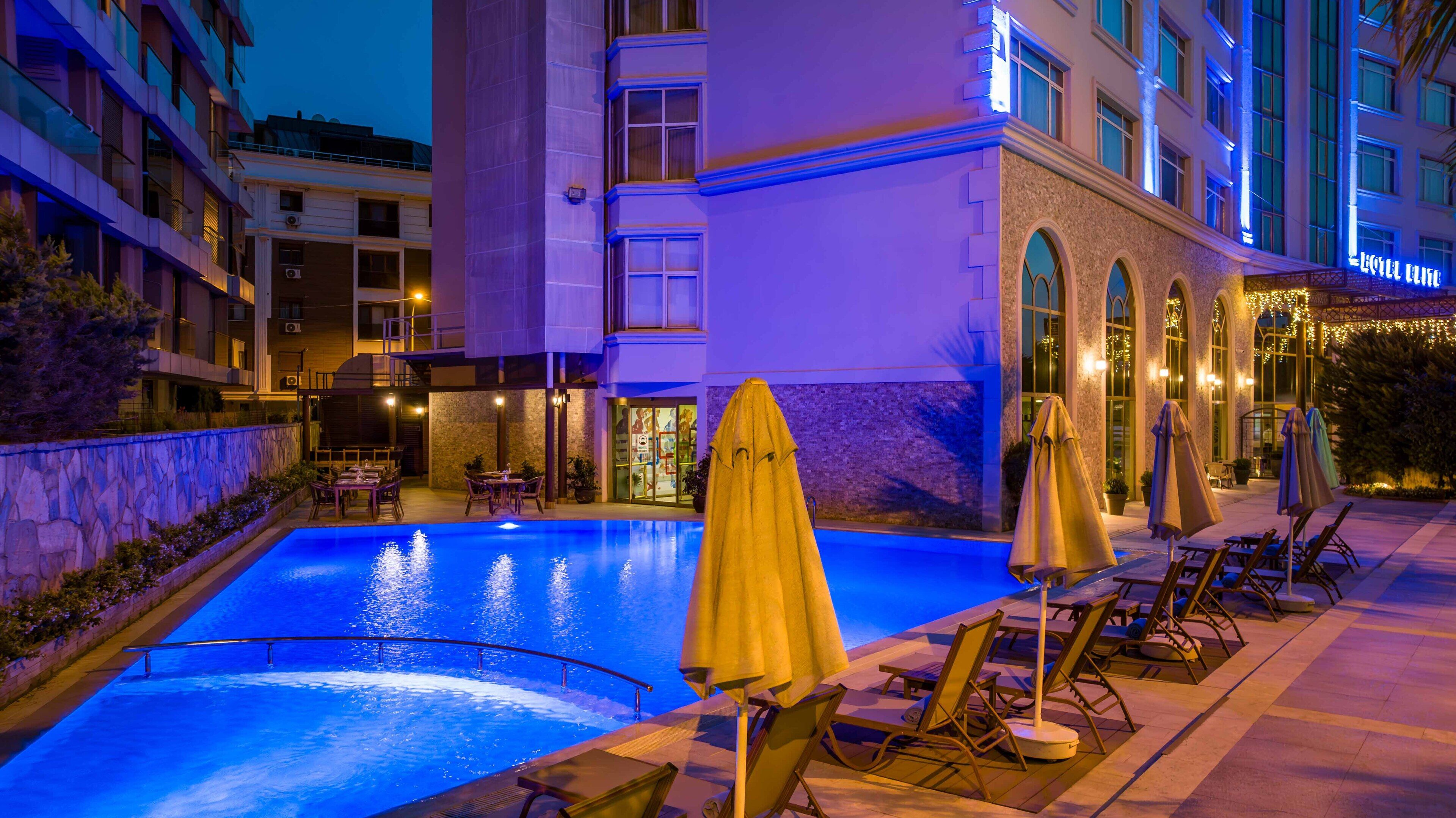 Un espace piscine d’hôtel avec des chaises longues et des parasols, entouré de bâtiments dont les fenêtres sont illuminées.