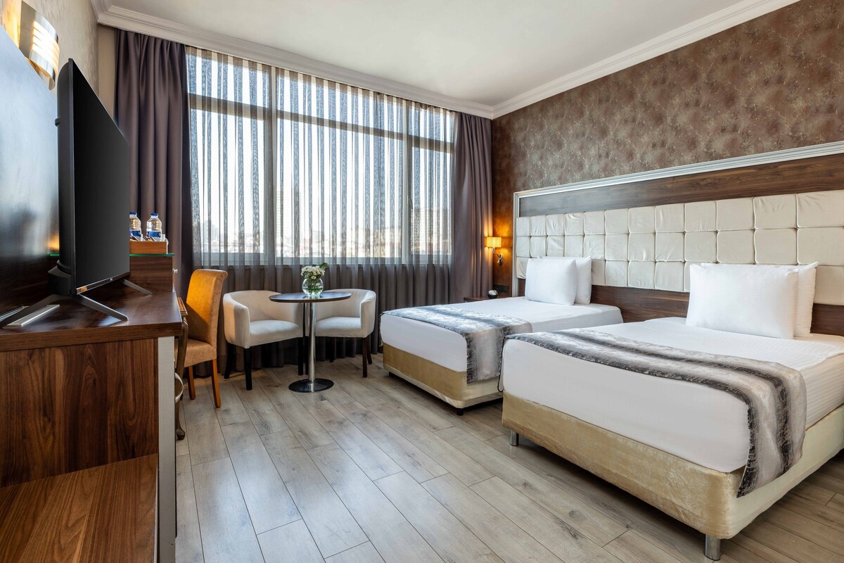 Ett hotellrum med två sängar, en TV, ett litet bord med stolar och ett fönster med gardiner.