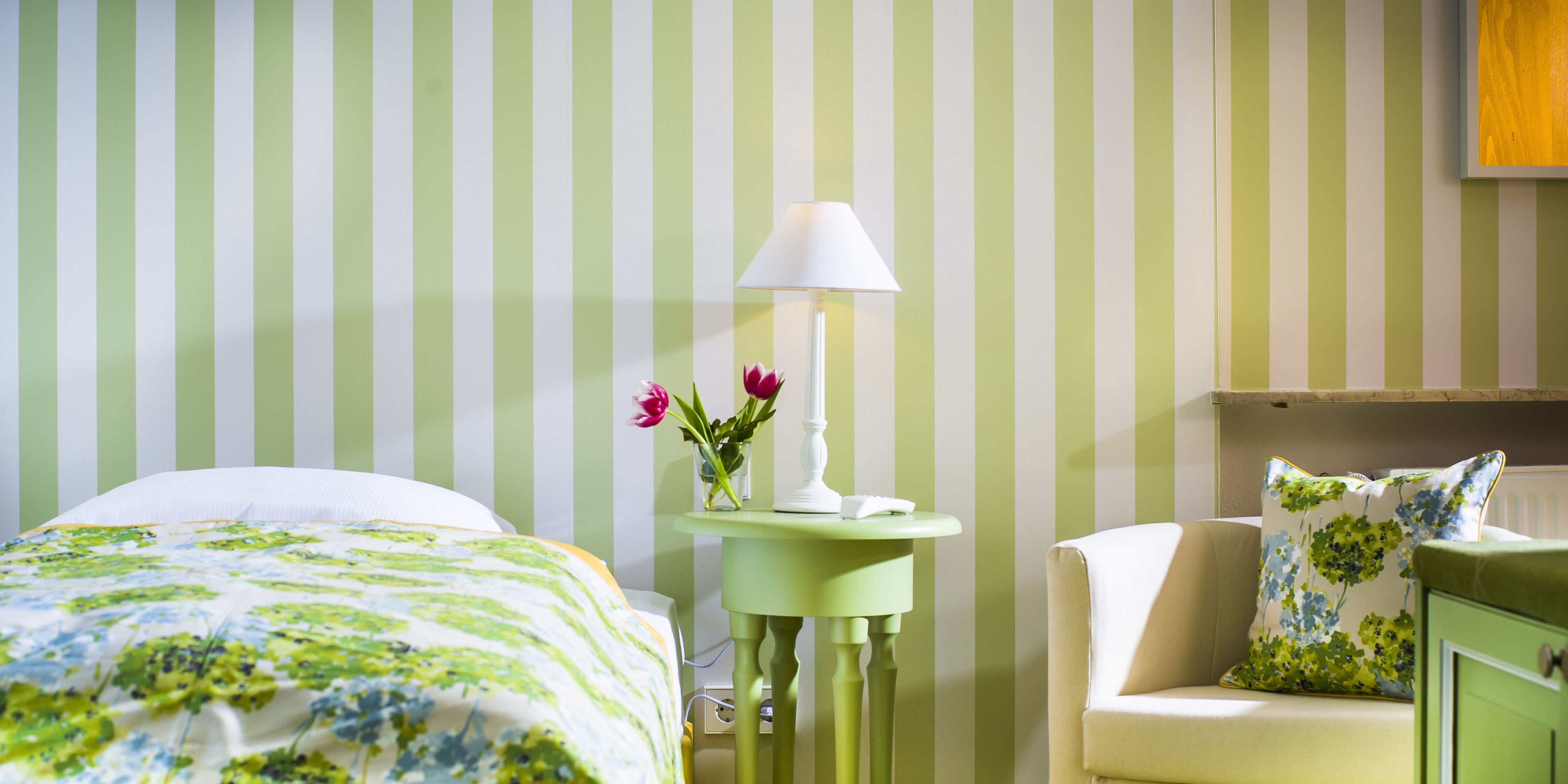 Camera d'albergo con un letto, un comodino con lampada, un vaso con fiori e una sedia con cuscino floreale.
