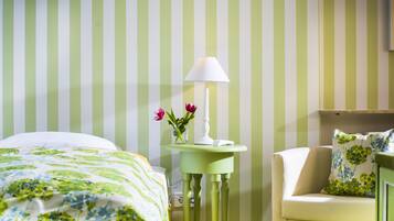 Camera d'albergo con un letto, un comodino con lampada, un vaso con fiori e una sedia con cuscino floreale.