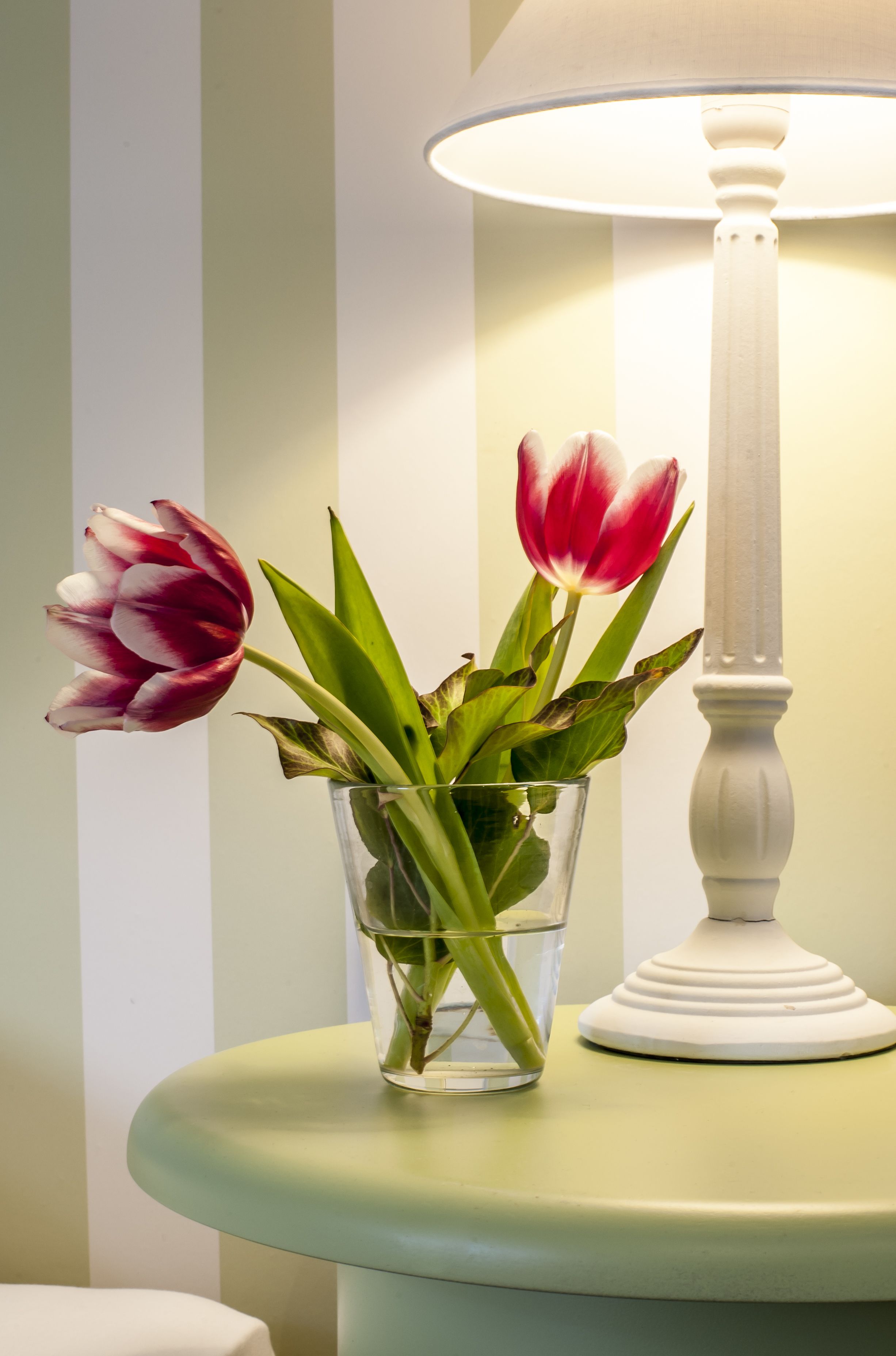 Un florero de vidrio transparente con agua y flores sobre una mesa, junto a una lámpara blanca.