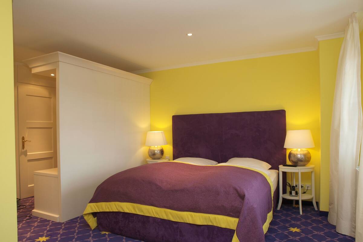 A hotel room with a bed, two bedside lamps, a wardrobe, and a bathroom with a shower curtain.