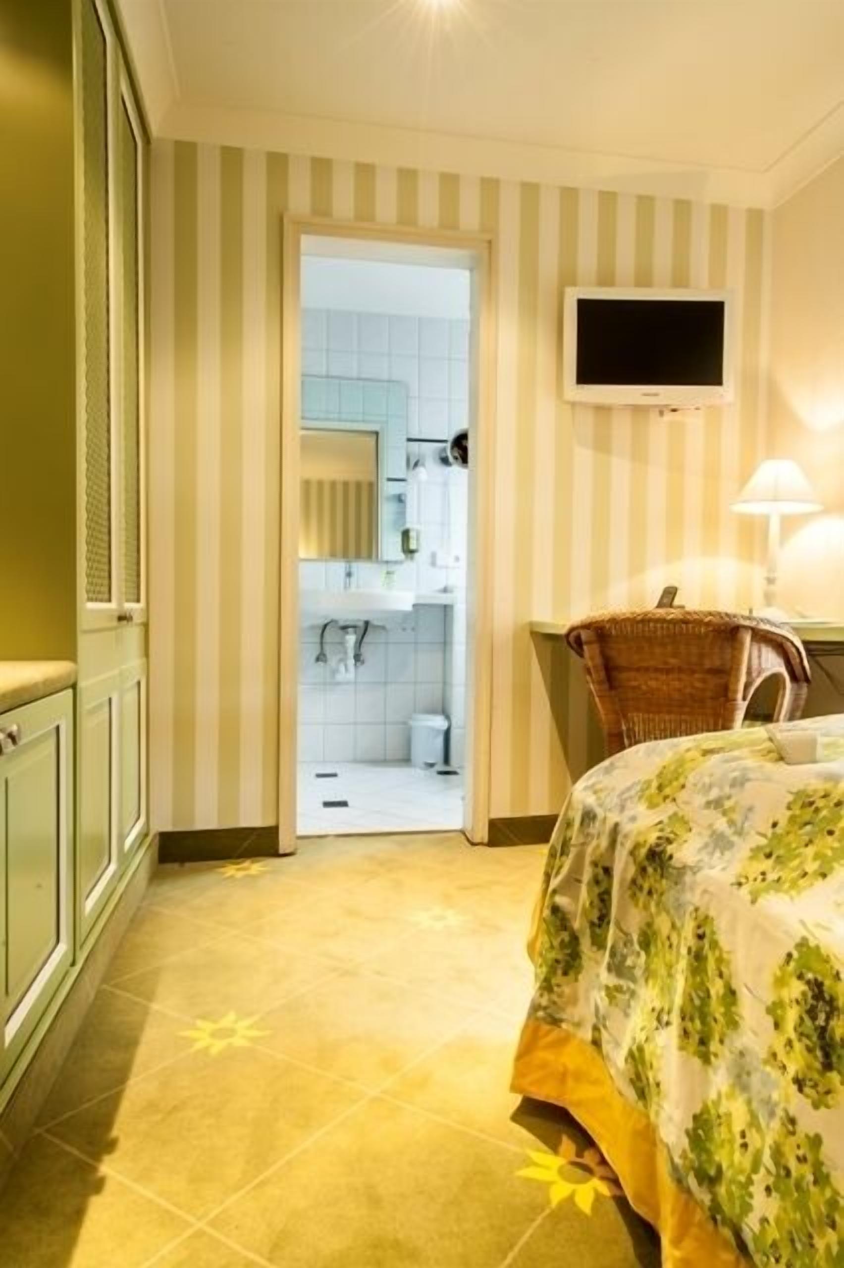 Habitación de hotel con cama, televisor montado en la pared, una silla de mimbre y baño visible a través de una puerta abierta.