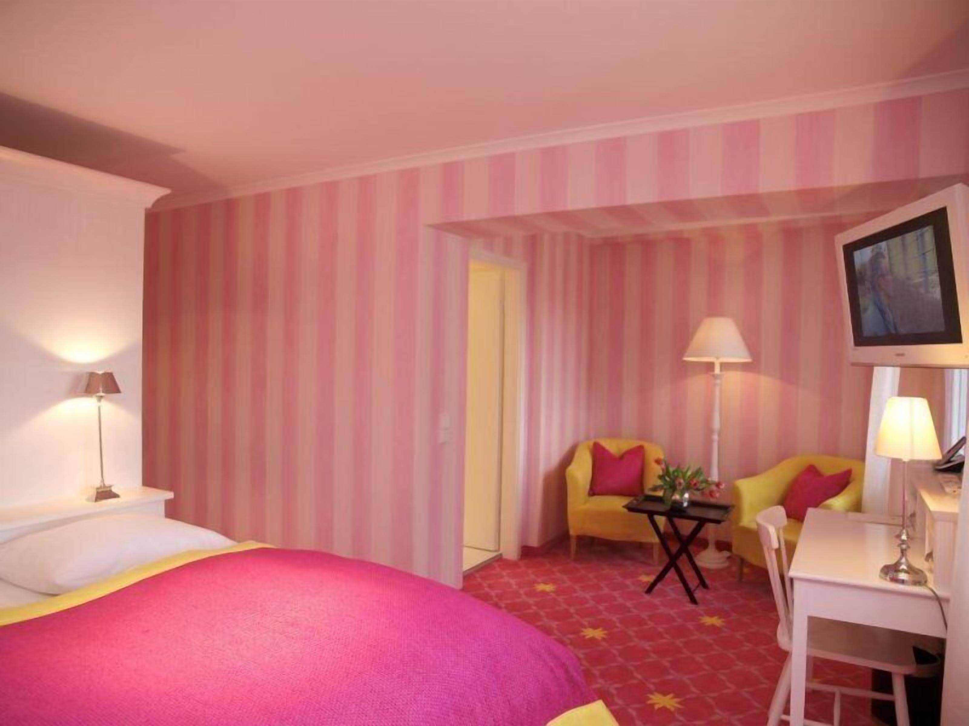 A hotel room with striped wallpaper, a bed, a desk, a chair, and a television.