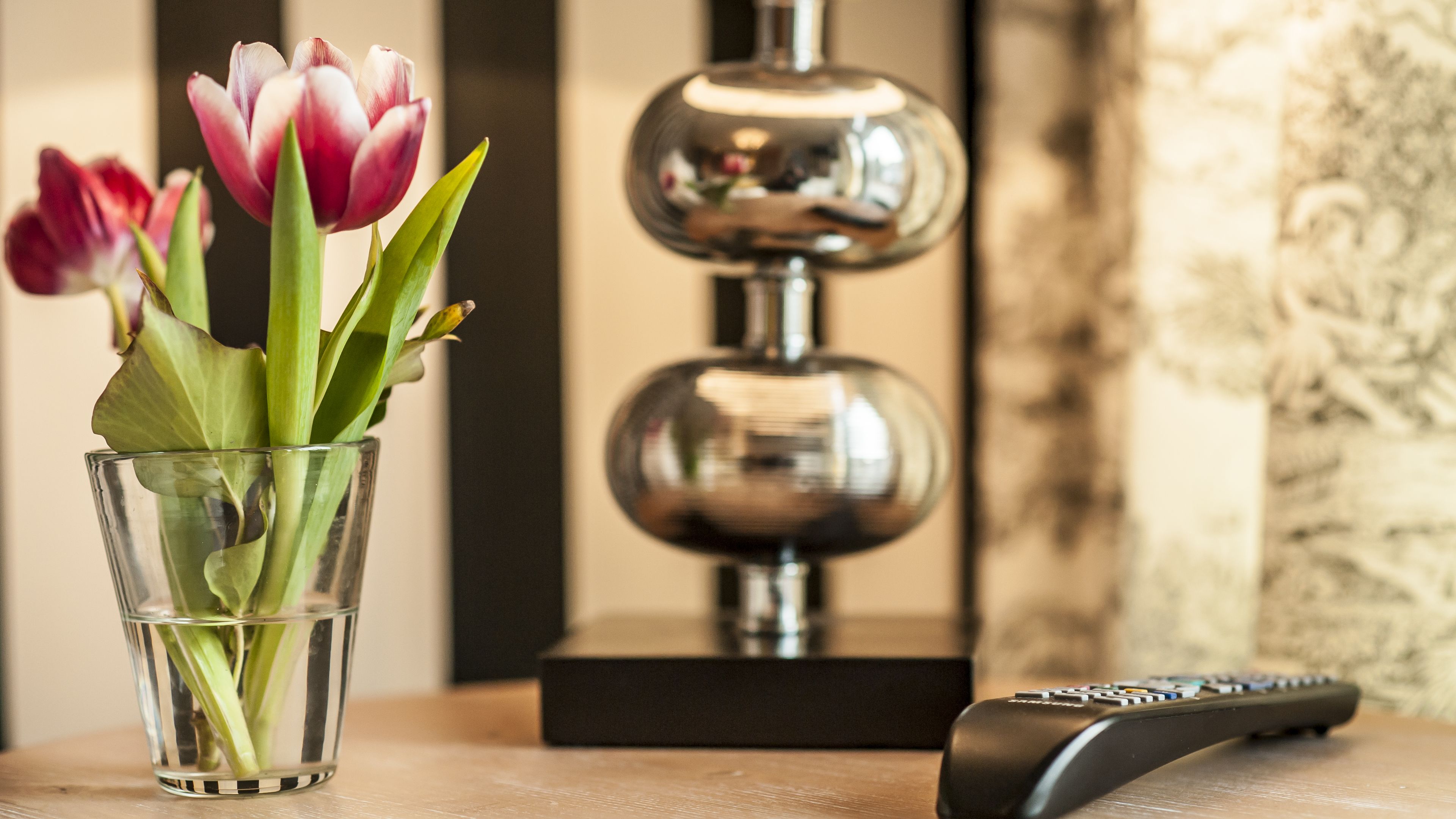 A glass vase with tulips on a wooden surface, a black remote control, and a decorative lamp in the background.