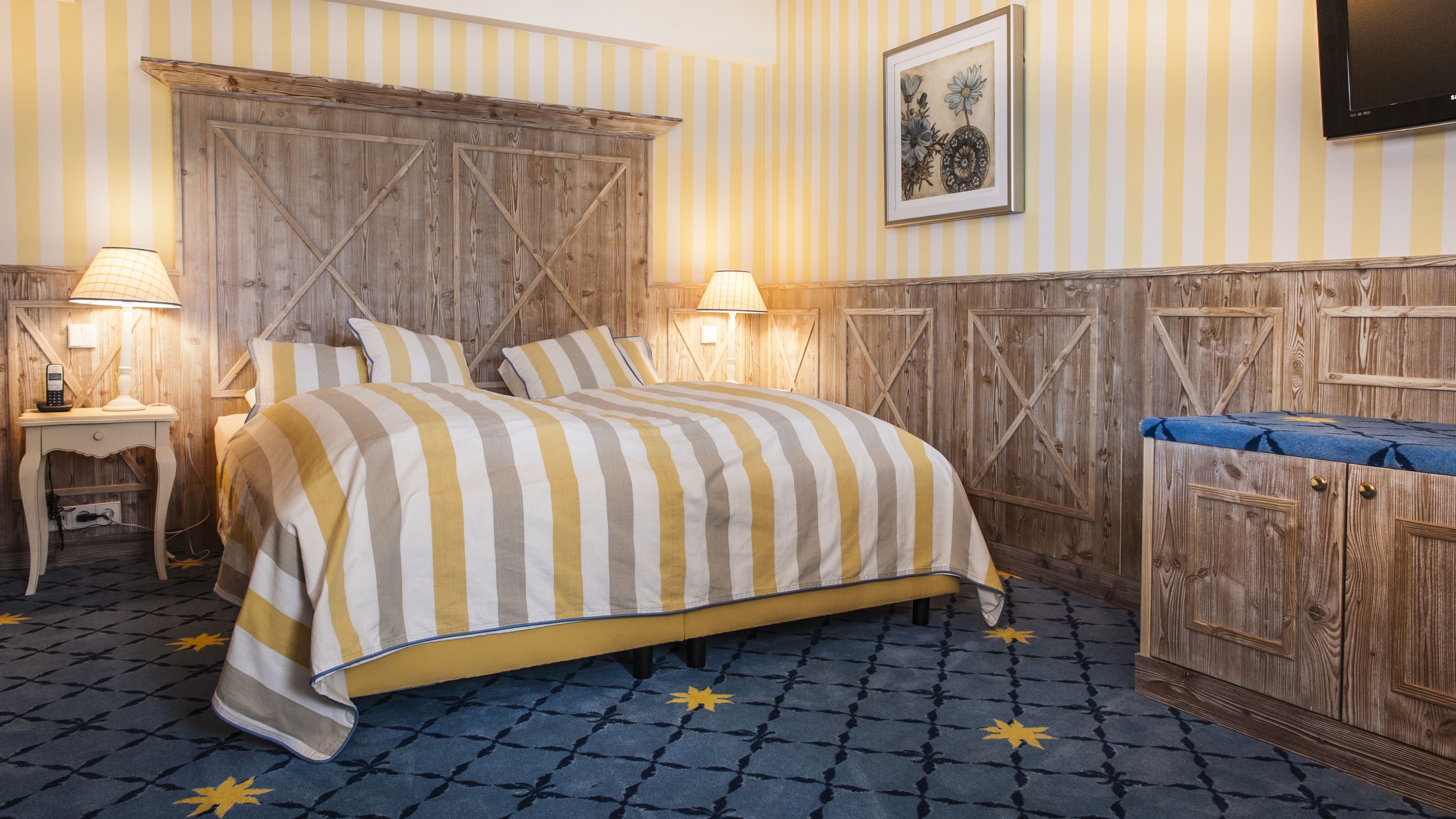 Een kamer met een groot bed, twee nachtkastjes met lampen, een houten hoofdeinde, een blauw kamerbreed tapijt en een aan de muur bevestigde televisie.