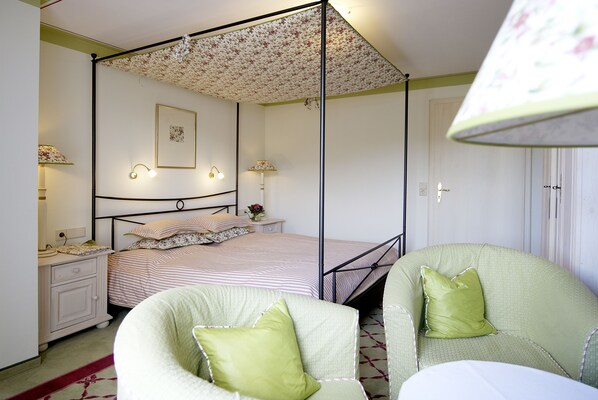 Un dormitorio con una cama con dosel, dos mesitas de noche con lámparas, un sillón verde y un dosel estampado.