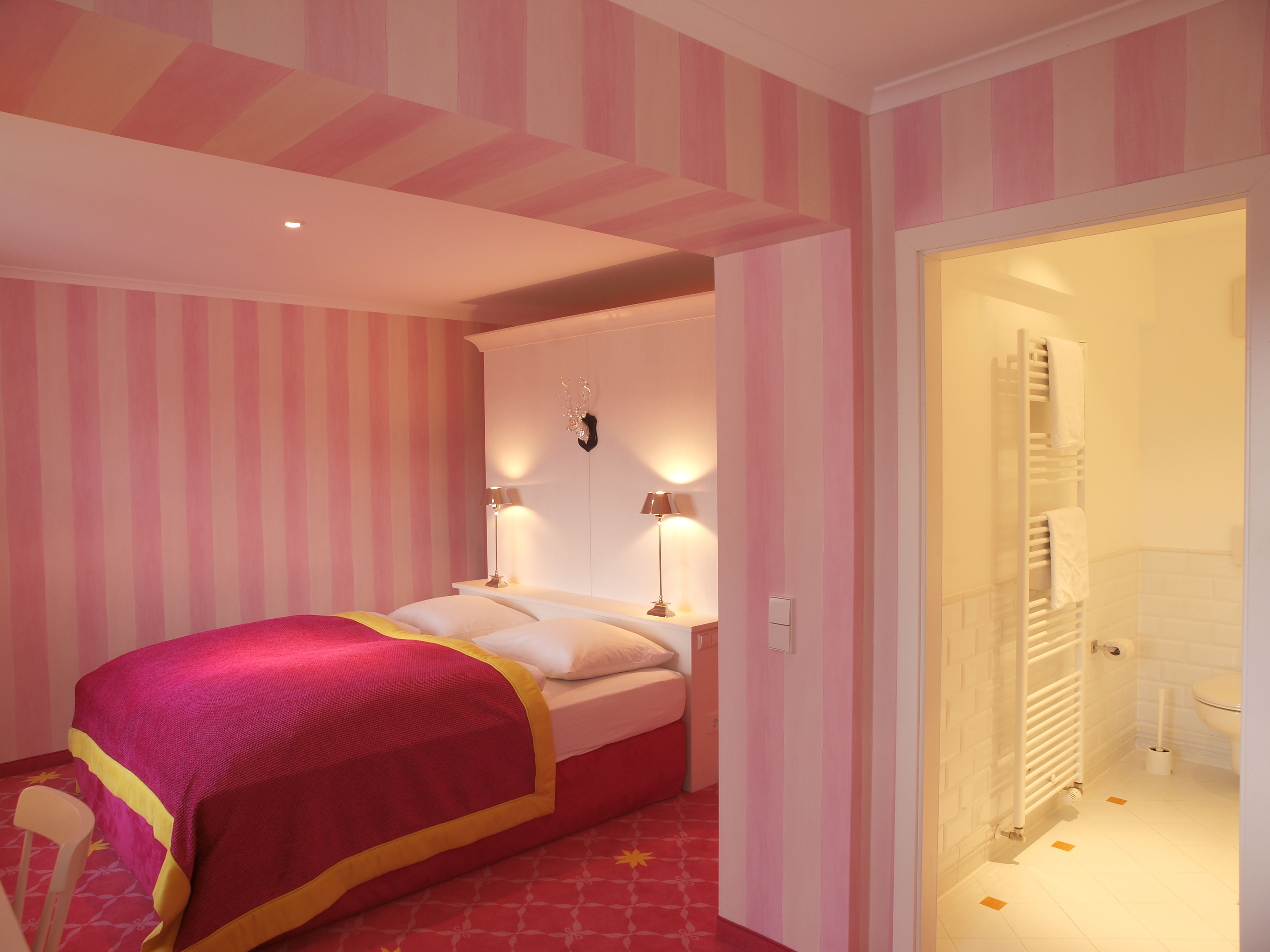 Un dormitorio con cama, lámparas de noche, cabecero y baño a la vista a través de una puerta abierta.