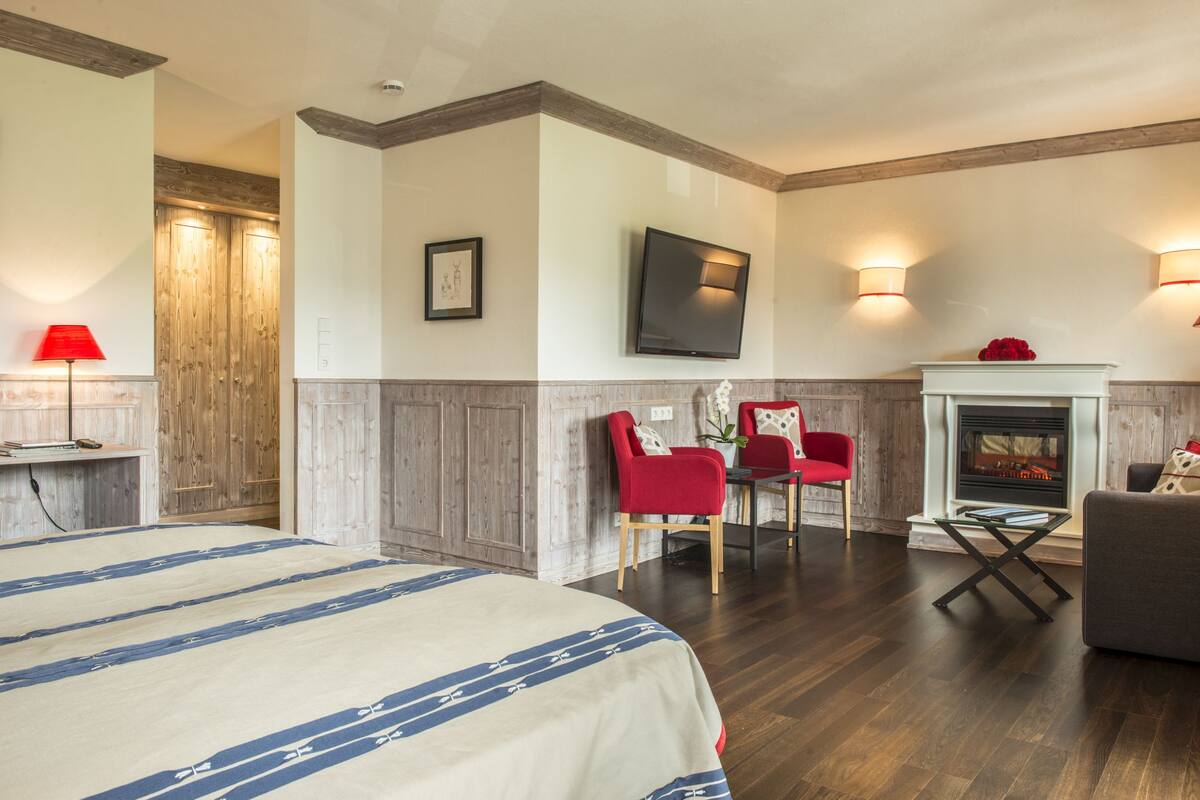 A hotel room with a bed, a fireplace, a TV, and a dining area with red chairs.