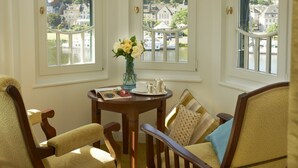 Une pièce avec vue sur une rivière et des bâtiments, une table ornée d’un vase de fleurs et deux fauteuils avec des coussins.