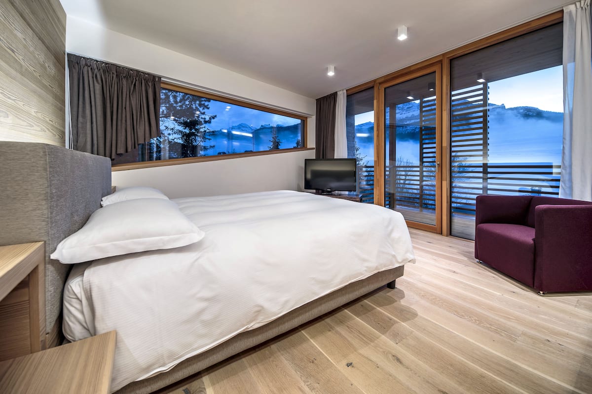 Habitación de hotel moderna con una cama grande, televisión y vistas a un lago y montañas a través de una puerta corrediza de vidrio.
