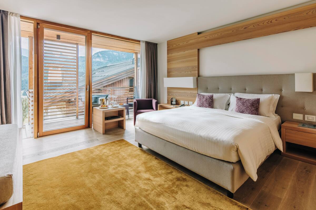 A modern bedroom with a large bed, a wooden headboard, a nightstand, a desk, and a chair. There is a sliding glass door leading to a balcony with a view of mountains.