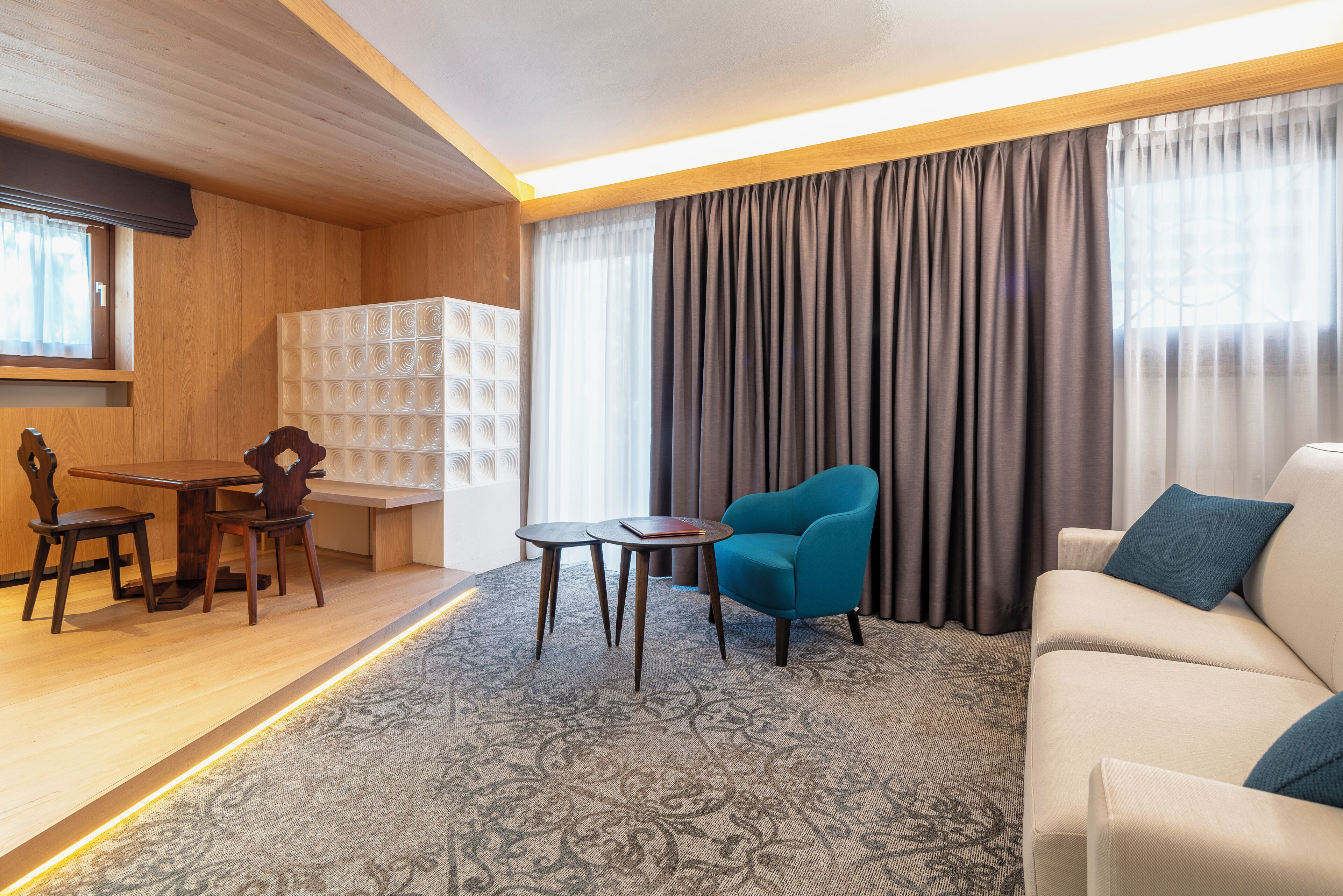 A modern hotel room with a wooden ceiling, a round table, a blue armchair, a sofa, and a patterned carpet.