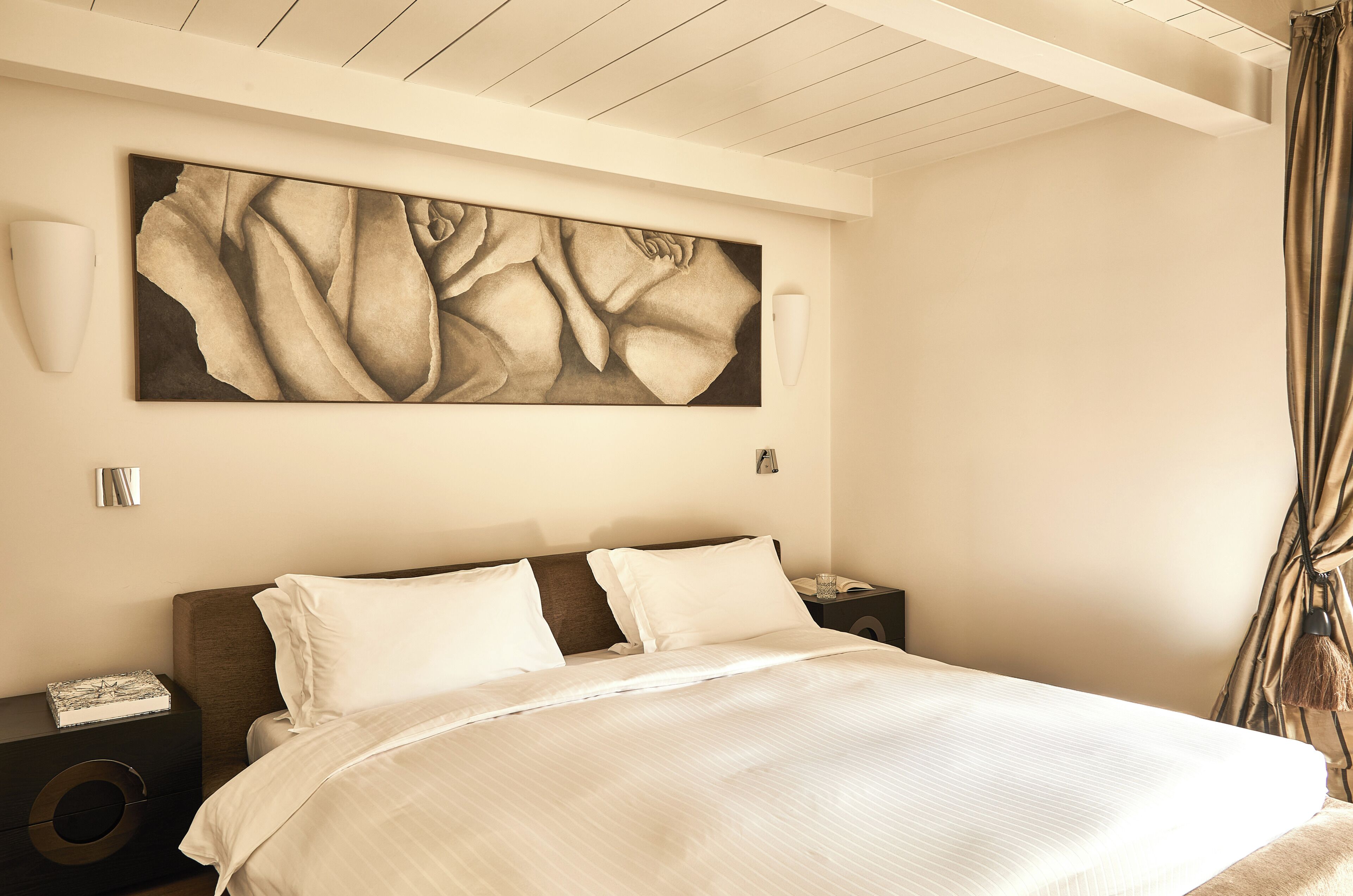 A hotel room with a large bed, a nightstand, a wall-mounted lamp, and a painting.