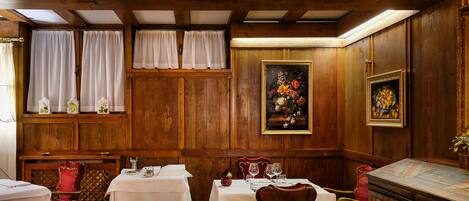 Comedor con paredes de paneles de madera, cortinas blancas y sillas tapizadas en rojo. Las mesas están dispuestas con manteles blancos y cubertería.