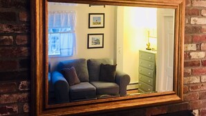Ein Spiegel mit Holzrahmen, der einen Raum mit einer Couch, einer Lampe und einer Kommode an einer Ziegelwand reflektiert.