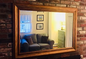 Un espejo con marco de madera que refleja una habitación con un sofá, una lámpara y un aparador frente a una pared de ladrillo.